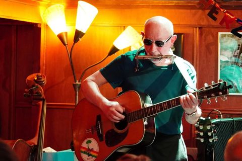 Un musicien joue de la guitare et de l'harmonica. En arrière-plan, on peut voir des lampes et d'autres instruments de musique.