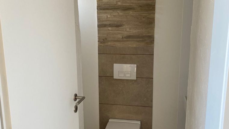 Ein modernes Badezimmer mit einer hellen Wand und einem eleganten, weißen WC. Der Fußboden ist mit grauen Fliesen ausgelegt.