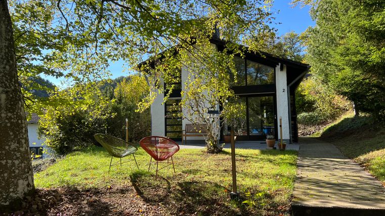 Een modern huis omringd door bomen en groen gras. Voor het huis staan twee gezellige stoelen.