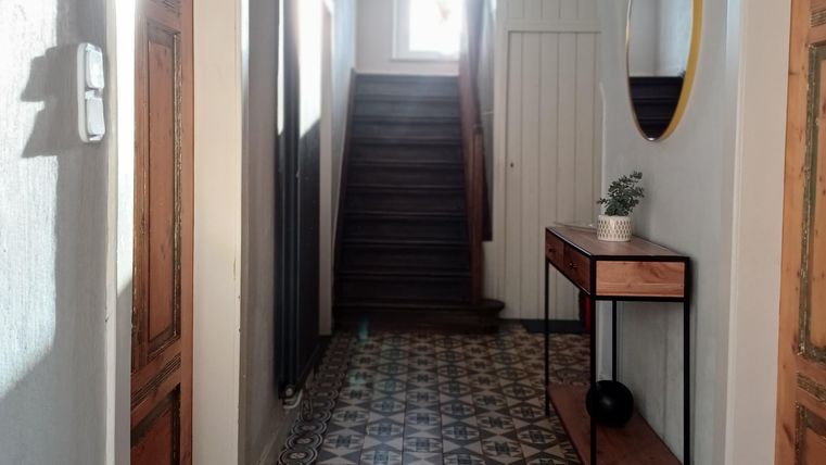 Een lichte gang met trap en grote ramen. Aan de muur hangt een ronde spiegel en een tafel met een plant.