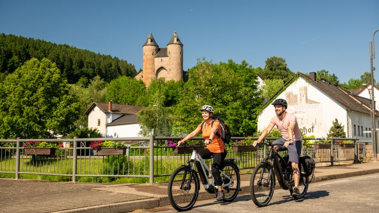 Zwei Radfahrer fahren auf einem Radweg entlang eines Flusses, im Hintergrund ist eine Burg zu sehen, am Ufer stehen Häuser