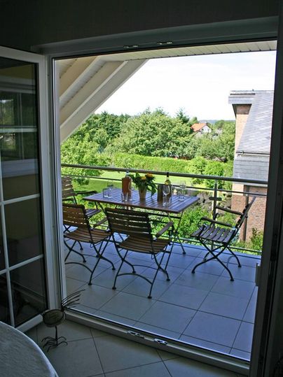 Een balkon met een tafel en stoelen, omringd door groene bomen. Door de grote glazen deur is een mooi uitzicht zichtbaar.