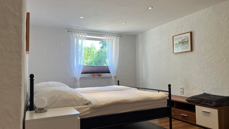 Een lichte slaapkamer met een tweepersoonsbed en een houten vloer. De ramen zijn ingericht met witte gordijnen en er is een nachtkastje en een commode.