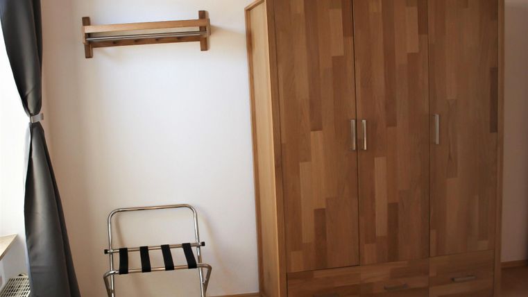 Ein einfaches Zimmer mit einem Holzkleiderschrank und einem Hocker. An der Wand hängt ein Holzregal.