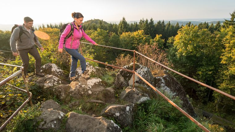 Zwei Wanderer auf einem felsigen Pfad mit Geländer, umgeben von Wald und Aussicht auf Hügel.