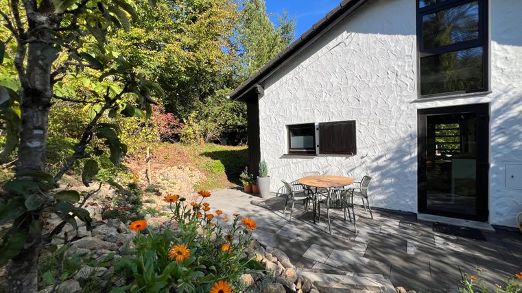 Een gezellig huis met een lichte gevel en grote ramen. Voor het huis staan een tafel en stoelen, evenals kleurrijke bloemen in een tuin.