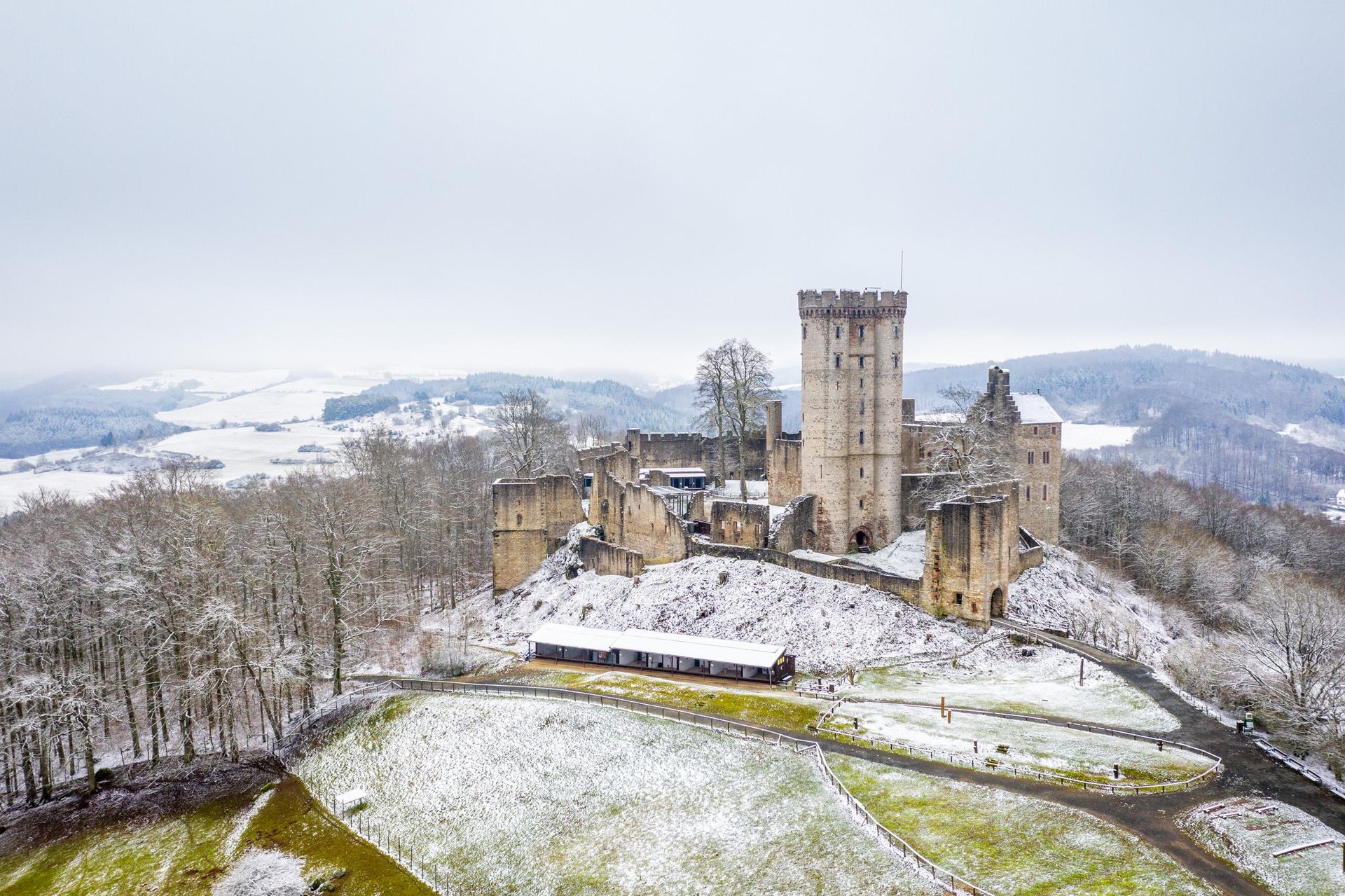 Eine massive Burg mit mehreren Türmen steht auf einer Felserhebung mitten in einer weitläufigen Wald- und Wiesenlandschaft im Winter. Die freien Wiesen sind leicht mit Schnee bedeckt.
