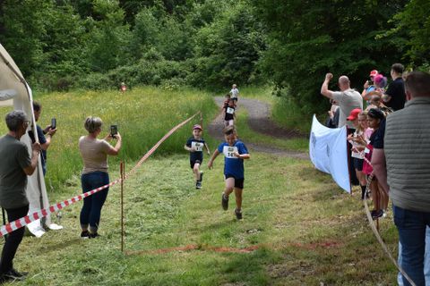 A group of children is taking part in a race while spectators stand at the sidelines taking photos. Several children are running on a field path, and some of the spectators are cheering them on.