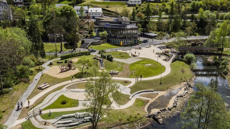 Luftaufnahme eines Kurparks mit geschwungenen Wegen, Spielplatz, Grünflächen und einem Fluss. Im Hintergrund sind Gebäude und Bäume zu sehen.
