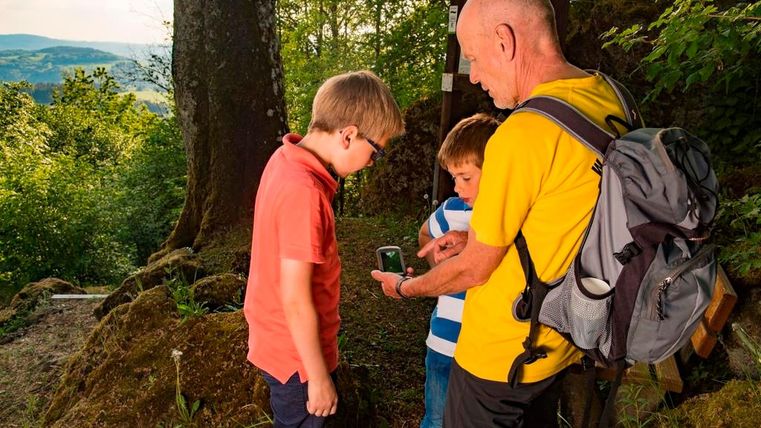 Zwei Kinder stehen mit einem Mann im Wald. Alle drei schauen auf ein GPS-Gerät.