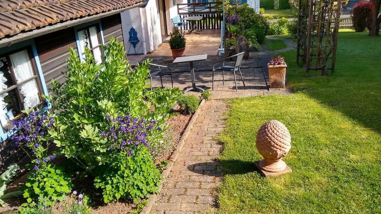 Ein gepflegter Garten mit bunten Blumen und einem gepflasterten Weg. Im Hintergrund sieht man eine Terrasse mit Stühlen und einen dekorativen Pflanzkübel.