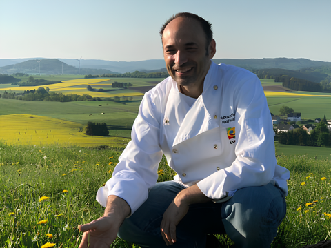 Ein lächelnder Koch kniet auf einer Wiese mit gelben Blumen. Im Hintergrund erstreckt sich eine malerische Landschaft mit sanften Hügeln und Feldern.