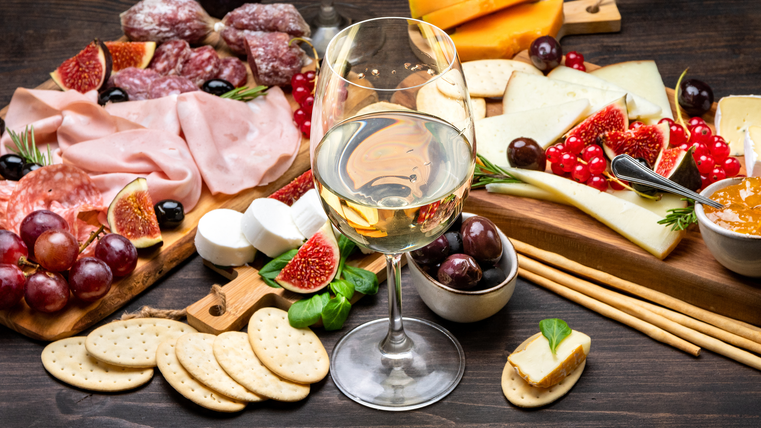 Auf einem dunklen Holzbrett liegen diverse Antipasti (Schinken, Salami, Käse, aufgeschnittene Feigen, Trauben, Cracker) und ein Glas Wein