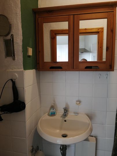 Een badkamer met een rond wasbak en een houten spiegel. Aan de muur hangt een haardroger en de tegels zijn licht.