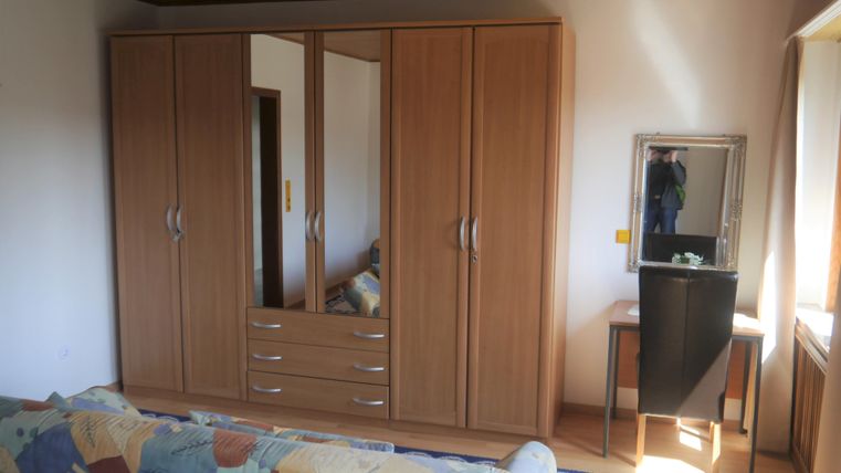 Ein moderner Kleiderschrank aus Holz mit Spiegeln und Schubladen. Im Hintergrund ist ein Schminktisch zu sehen, sowie ein gemütliches Sofa.