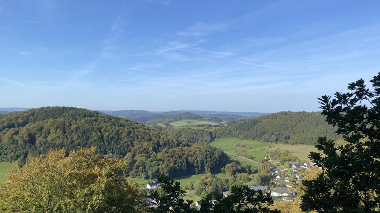 Panoramablick vom Mühlberg auf bewaldete Hügel und ein Dorf im Tal unter klarem, blauem Himmel.
