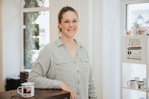 Eine Frau steht hinter einem Tresen und lächelt in die Kamera. Hinter ihr einige Fenster und Regale mit regionalen Produkten.