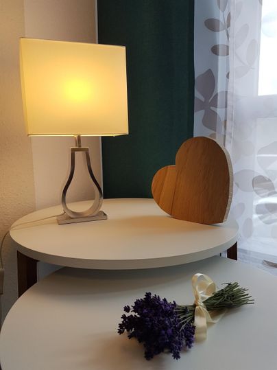 Eine Tischlampe leuchtet sanft auf einem runden Tisch. Daneben liegt ein Herz aus Holz und ein Sträußchen Lavendel.