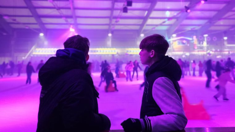 Deux personnes se tiennent au bord d'une patinoire et observent les patineurs. Des lumières colorées créent une atmosphère festive.