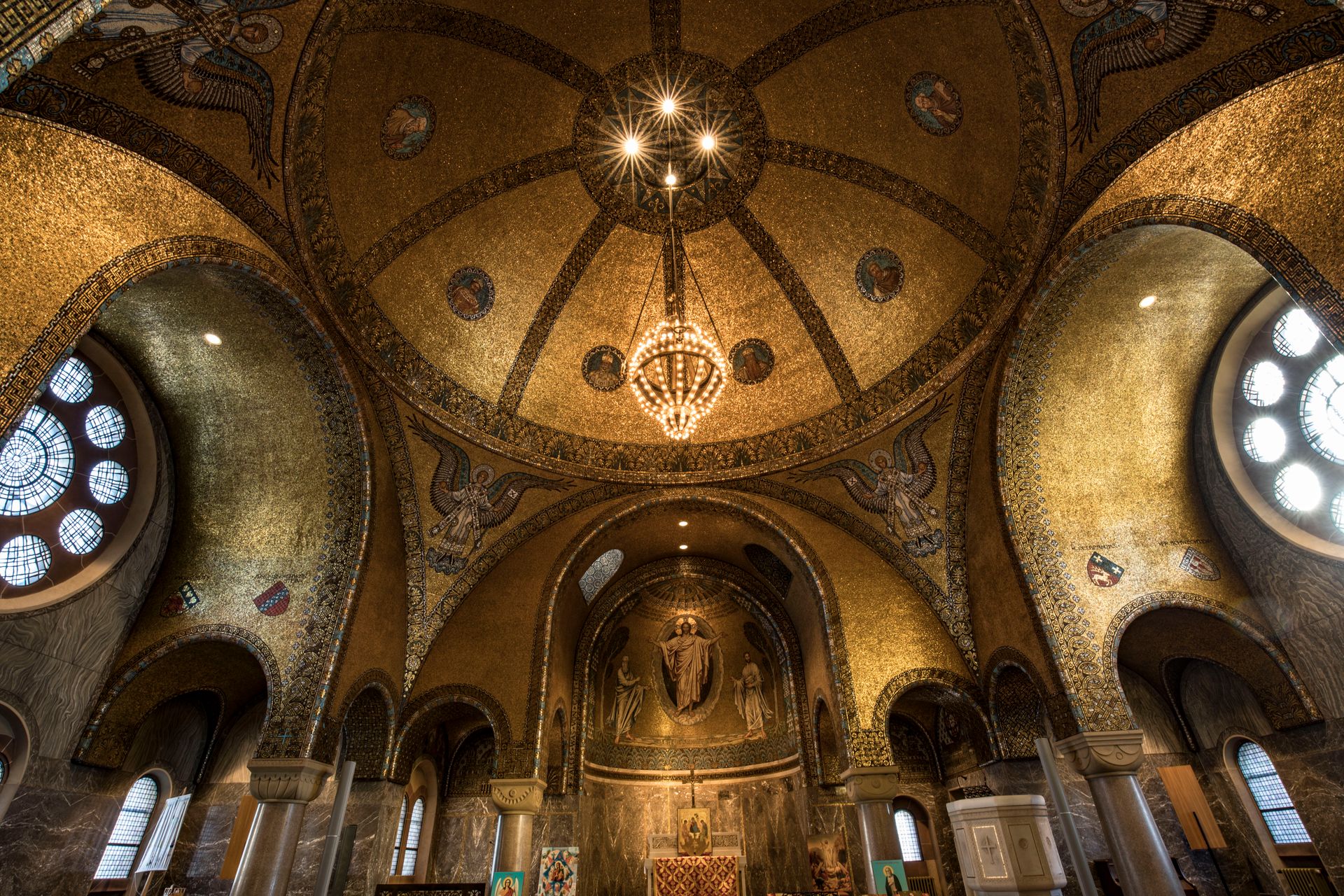 Innenraum einer mit unzähligen goldenen Mosaiksteinen verzierten Kirche mit großen Seitenfenstern, durch die Licht hineinfällt.