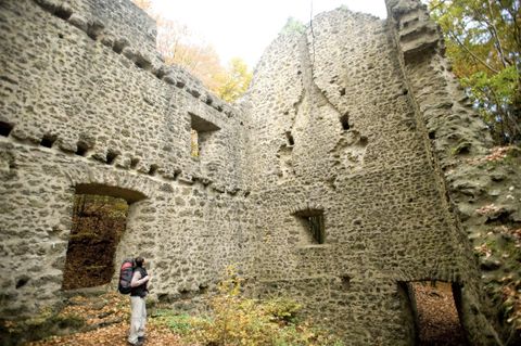Ein Wanderer steht vor den Ruinen einer alten Steinmauer im Wald. Die Mauer hat Fensteröffnungen und ist von Herbstlaub umgeben.
