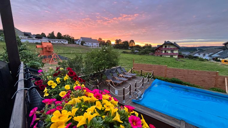 Eine blühende Blumenlandschaft mit bunten Pflanzen im Vordergrund. Im Hintergrund sieht man ein ruhiges Landhaus und einen Sonnenuntergang am Horizont.