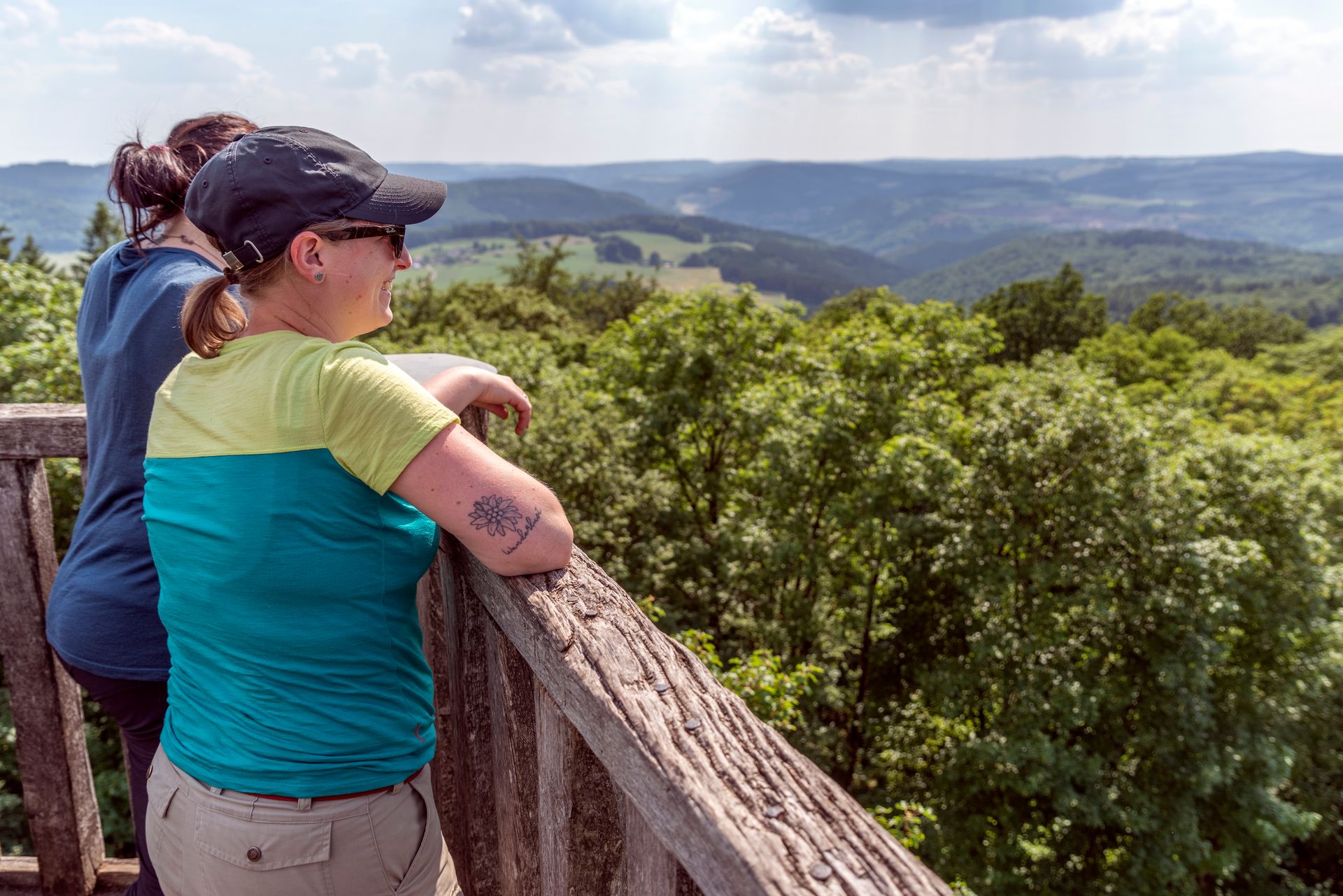 Zwei Personen in Outdoorkleidung lehnen am Geländer eines hözernen Turms und schauen in die weitläufige Waldlandschaft.
