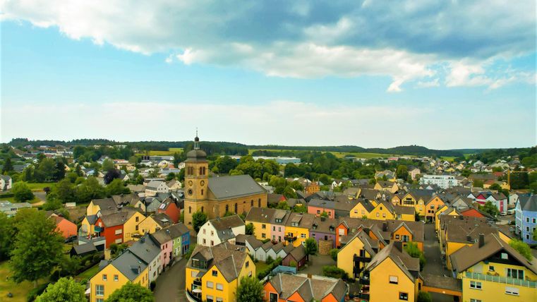 Eine malerische Ansicht eines kleinen Dorfes mit bunten Häusern und einer Kirche. Im Hintergrund sind grüne Hügel und ein klarer Himmel zu sehen.