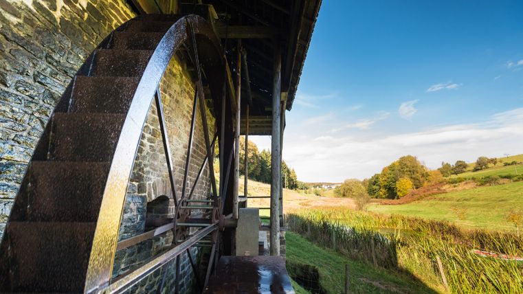 A large mill wheel on an old stone wall, surrounded by a green landscape and blue sky.