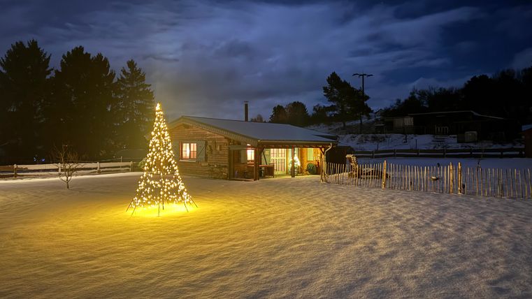 Ein gemütliches Haus im Schnee bei Nacht, umgeben von einem glitzernden Lichtbaum. Der Himmel ist bewölkt und die Atmosphäre ist ruhig und festlich.