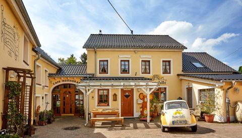 Ein charmantes Café-Restaurant mit gelben Wänden und einem einladenden Eingang. Im Vordergrund steht ein klassisches Auto und die Umgebung ist freundlich und einladend.