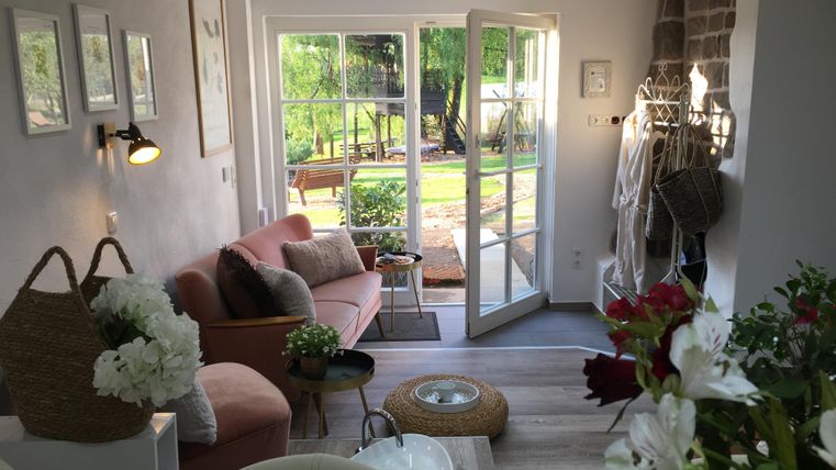 Un espace de vie confortable avec un canapé rose et de grandes fenêtres donnant sur une vue de jardin. Des arrangements floraux et une atmosphère lumineuse et accueillante rendent la pièce chaleureuse et amicale.