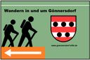 Schild mit Wanderern, Wappen und Pfeil nach links.