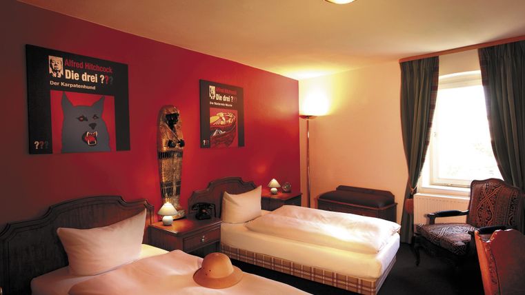 Une chambre confortable avec deux lits simples et un mur rouge.
Des tableaux sont accrochés aux murs et il y a une fenêtre avec des rideaux.
