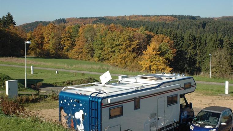 Wohnmobil und Auto auf einem Stellplatz mit herbstlichem Wald im Hintergrund. Der Himmel ist klar und blau.