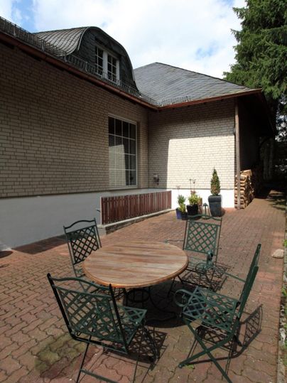 Ein gemütlicher Außenbereich mit einem runden Tisch und vier Stühlen aus Metall. Im Hintergrund ist ein Haus und einige Pflanzen zu sehen.