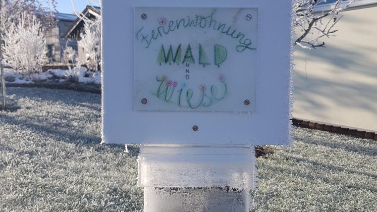 Ein beschrifteter Briefkasten steht in einer frostigen Umgebung. Im Hintergrund erkennt man gefrostete Bäume und ein Wohnhaus.