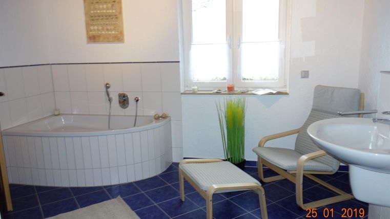 Ein Badezimmer mit einer Badewanne, einem Waschbecken und einem bequemem Stuhl. Große Fenster lassen viel Licht herein und es gibt grünes Dekor.