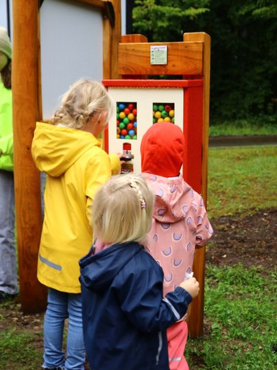 Drei Kinder in Regekjacken stehen dicht beieinander vor dem Kugelautomat und kaufen Kugeln für die Waldkugelbahn.