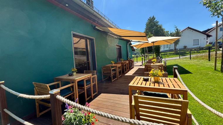 Ein gemütlicher Außenbereich mit Holztischen und Schirmen. Die Sonne scheint auf das grüne Gebäude und den gepflegten Rasen.