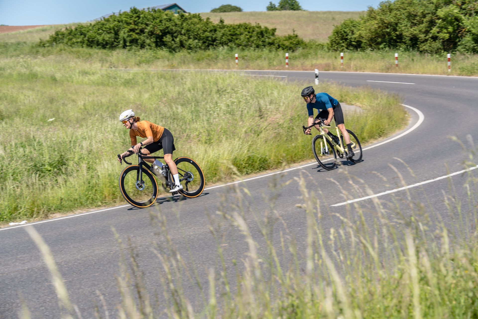 Zwei Radfahrer auf Rennrädern fahren durch eine steile Kurve auf einer Landstraße umgeben von Wiesen.