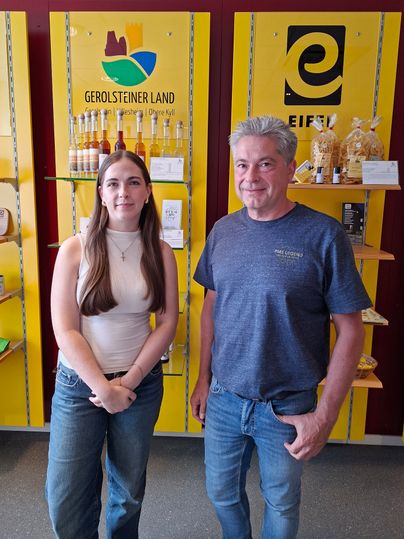Ein Mann und eine junge Frau stehen nebeneinander vor eine Shop-Wand und lächeln in die Kamera.
