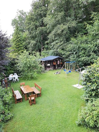 Ein schöner Garten mit einer grünen Wiese, einem Holzspielplatz und einem überdachten Bereich. Es gibt auch Möbel wie Tische und Stühle zum Entspannen.