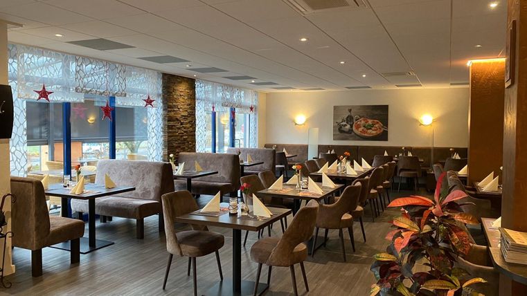 Ein gemütliches Restaurant mit gedeckten Tischen, braunen Stühlen und Dekoration. An den Fenstern hängen rote Sterne, ein Bild zeigt eine Pizza.