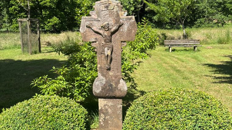 Stone cross with figure of Jesus in a garden with bushes and trees in the background.