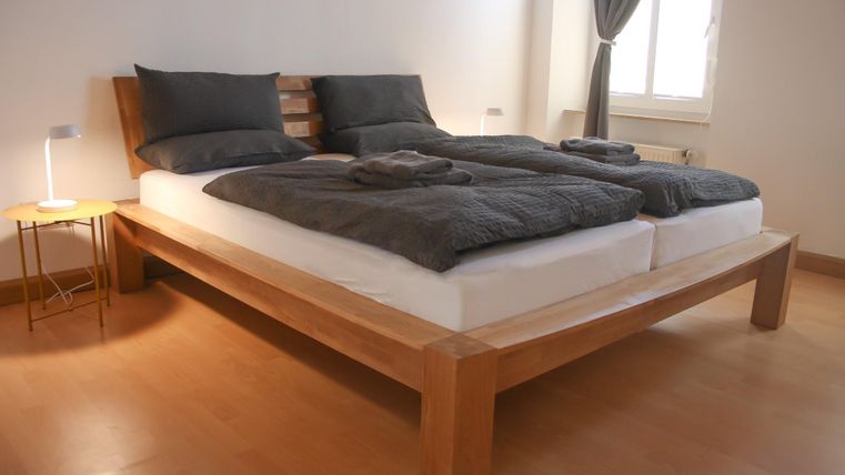 Ein modernes Schlafzimmer mit einem Holzbett und grauer Bettwäsche. Das Zimmer ist hell und freundlich, mit einem Tisch und einem Fenster.