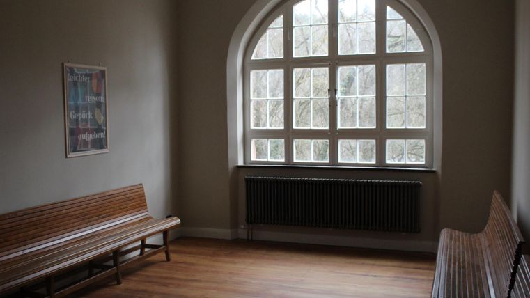 Leere Wartehalle mit Holzbänken, großem Fenster und einem Plakat an der Wand. Holzboden und Heizkörper unter dem Fenster.