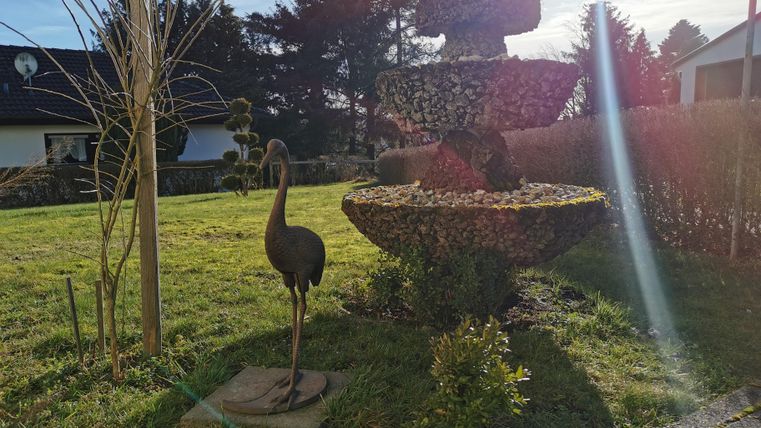 A well-maintained garden with an ornate pot of trimmed plants and a sculpture of a heron-like bird. In the background, additional green areas and trees are visible.