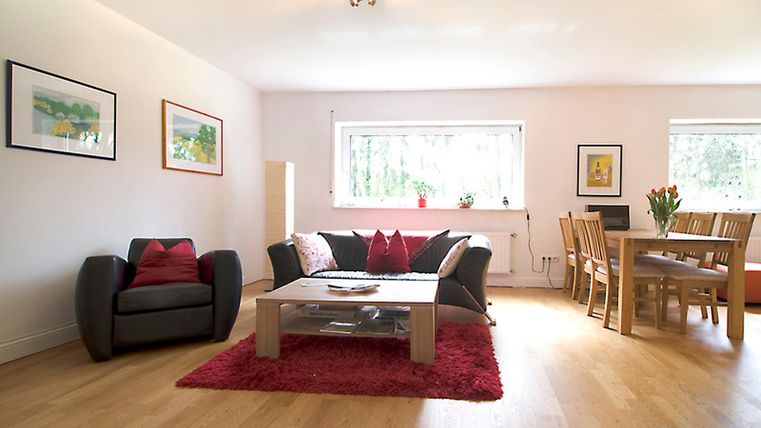 Ein modernes Wohnzimmer mit einem gemütlichen Sofa und einem Esstisch. Große Fenster lassen viel Licht herein und der Boden ist aus hellem Holz.