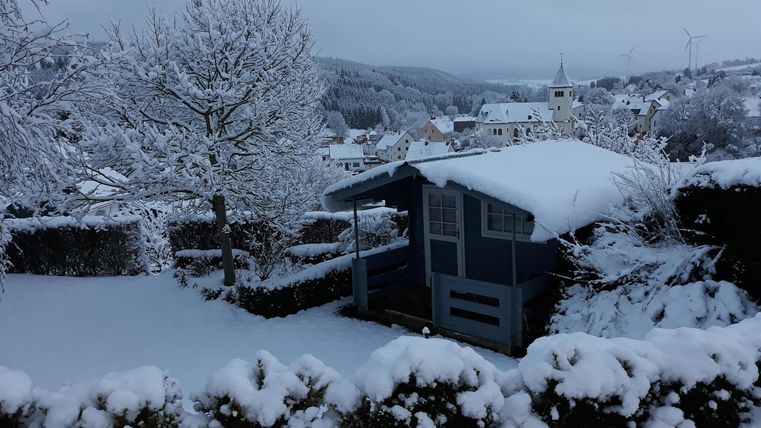 Eine verschneite Landschaft mit einem kleinen Gartenhaus und schneebedeckten Bäumen. Der Himmel ist grau und es herrscht Winterstimmung.